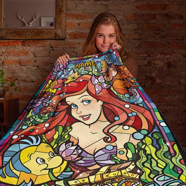 Puede incluir: Una manta rectangular con una vibrante ilustraci&oacute;n estilo vidriera de una sirena de pelo rojo y un pez amarillo. El dise&ntilde;o incluye plantas acu&aacute;ticas, burbujas y un cofre del tesoro, con la palabra NAME impresa en la parte superior.