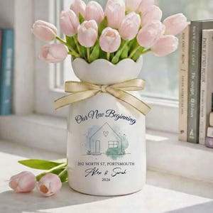 May include: A white ceramic vase with a scalloped top, tied with a gold ribbon, holds a bouquet of pink tulips. The vase is decorated with a house illustration and the text "Our New Beginning", "202 North St, Portsmouth", "Alex & Sarah", and "2026".