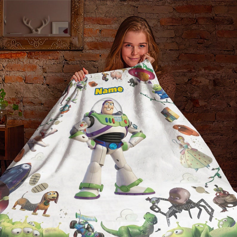 Puede incluir: Una manta de forro polar blanca con un gr&aacute;fico central grande de Buzz Lightyear, la palabra Name en letras amarillas y varias ilustraciones de personajes de Toy Story, incluyendo un dinosaurio, un perro y alien&iacute;genas verdes.