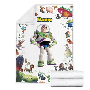 Puede incluir: Una manta de forro polar blanca con un gr&aacute;fico central grande de Buzz Lightyear, la palabra Name en texto amarillo y varias ilustraciones de personajes de Toy Story, incluyendo un cerdo, alien&iacute;genas, un perro y una nave espacial Pizza Planet.