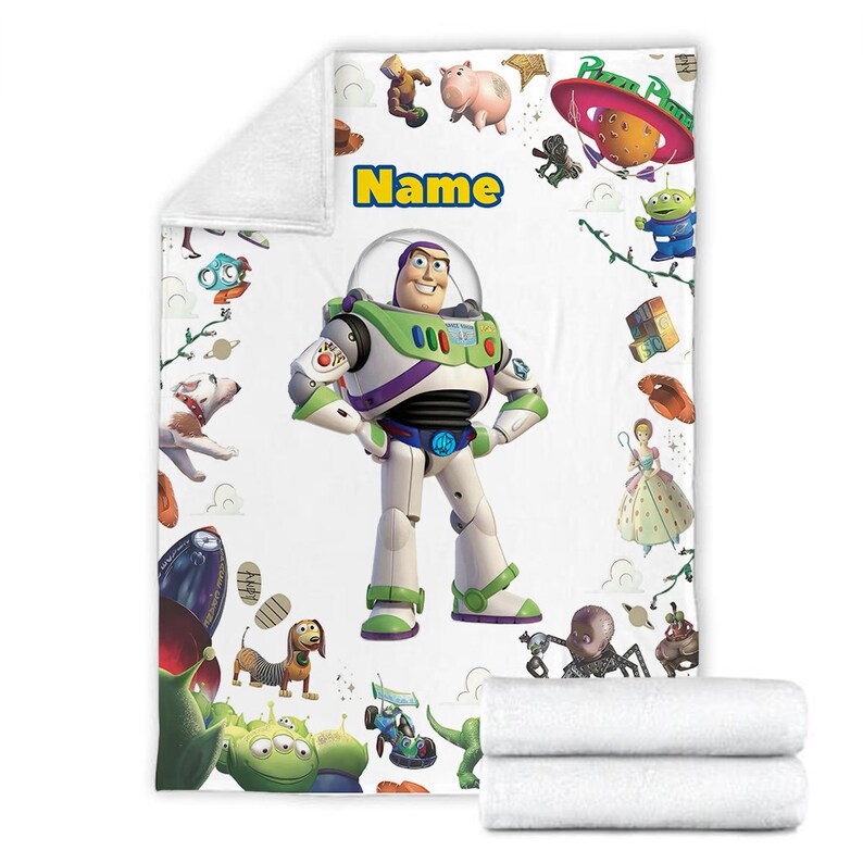 Puede incluir: Una manta de forro polar blanca con un gr&aacute;fico central grande de Buzz Lightyear, la palabra Name en texto amarillo y varias ilustraciones de personajes de Toy Story, incluyendo un cerdo, alien&iacute;genas, un perro y una nave espacial Pizza Planet.
