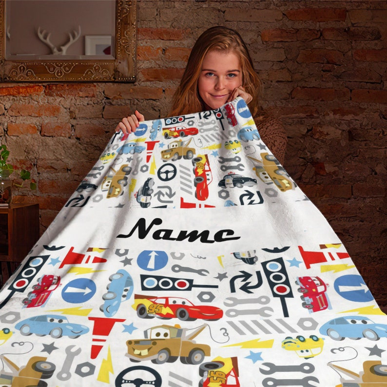 May include: A white fleece blanket featuring a repeating pattern of cartoon cars, traffic lights, road signs, and tools. The blanket includes a central horizontal band with the word Name printed in black script lettering across the centre.