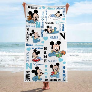 May include: A white beach towel with a blue and black Mickey Mouse design. The towel features multiple images of Mickey Mouse in different poses, along with the word "Name" repeated throughout the design.