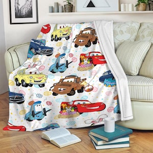 May include: A white fleece blanket with a colorful pattern of cartoon cars and Easter eggs. The cars are red, blue, yellow, and brown. The eggs are yellow, blue, pink, and green.