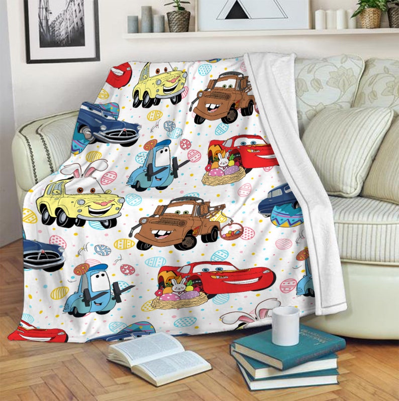 May include: A white fleece blanket with a colorful pattern of cartoon cars and Easter eggs. The cars are red, blue, yellow, and brown. The eggs are yellow, blue, pink, and green.