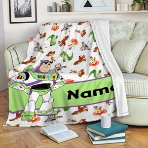 May include: A white fleece blanket featuring a repeating pattern of Toy Story characters including Buzz Lightyear, Woody, Jessie, and Rex. A large Buzz Lightyear figure stands on the left, next to a wide lime green horizontal band with the text Name.