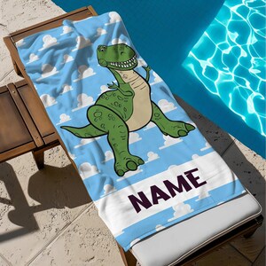 May include: A beach towel with a cartoon dinosaur design. The towel has a light blue sky background with white clouds and a green dinosaur. The word "NAME" is printed in dark purple across the bottom.