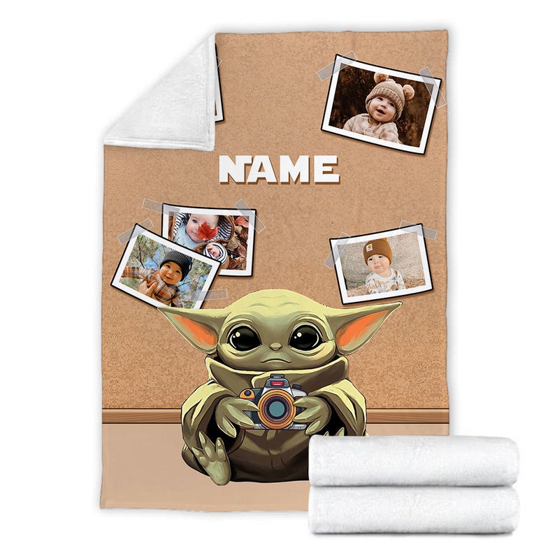 May include: A brown and tan fleece blanket with a cartoon image of a green alien holding a camera. The blanket has a corkboard background with photos of babies pinned to it. The text "NAME" is printed on the blanket.