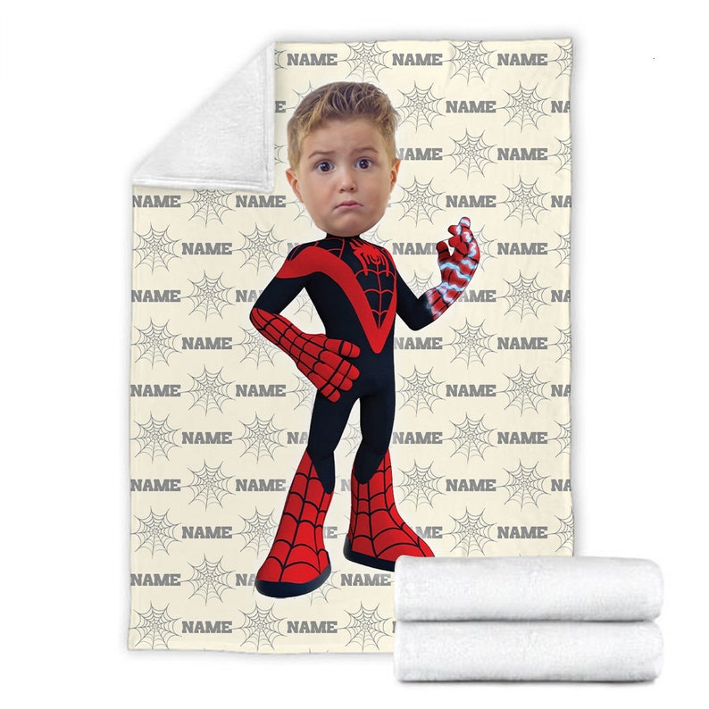 May include: A rectangular fleece blanket featuring a repeating pattern of grey spiderwebs and the word NAME in black text. Centered is a cartoon-style character wearing a black and red superhero suit with a child's face and one glowing hand.