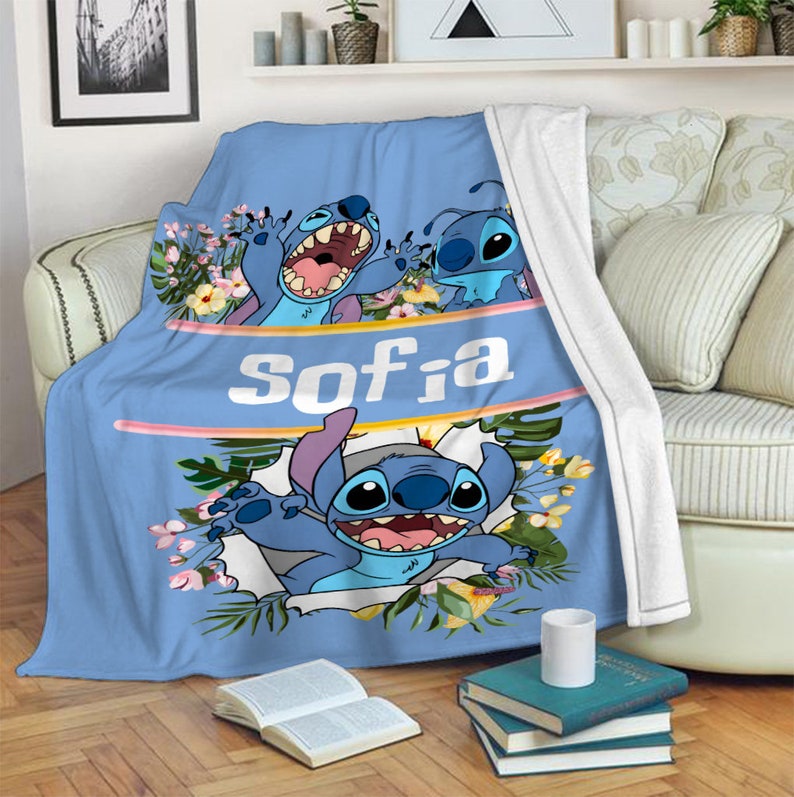 May include: A light blue fleece throw blanket featuring the character Stitch from Lilo & Stitch, surrounded by tropical flowers and green leaves. The name Sofia is printed in large white letters across a horizontal band in the center of the blanket.