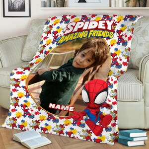 K&ouml;nnte beinhalten: Eine wei&szlig;e Fleecedecke mit einem wiederkehrenden Muster aus Spidey and His Amazing Friends-Figuren, gelben Sternen und roten Netzgrafiken. In der Mitte befindet sich ein gro&szlig;er ovaler Rahmen mit einem Kinderfoto und dem Wort NAME.