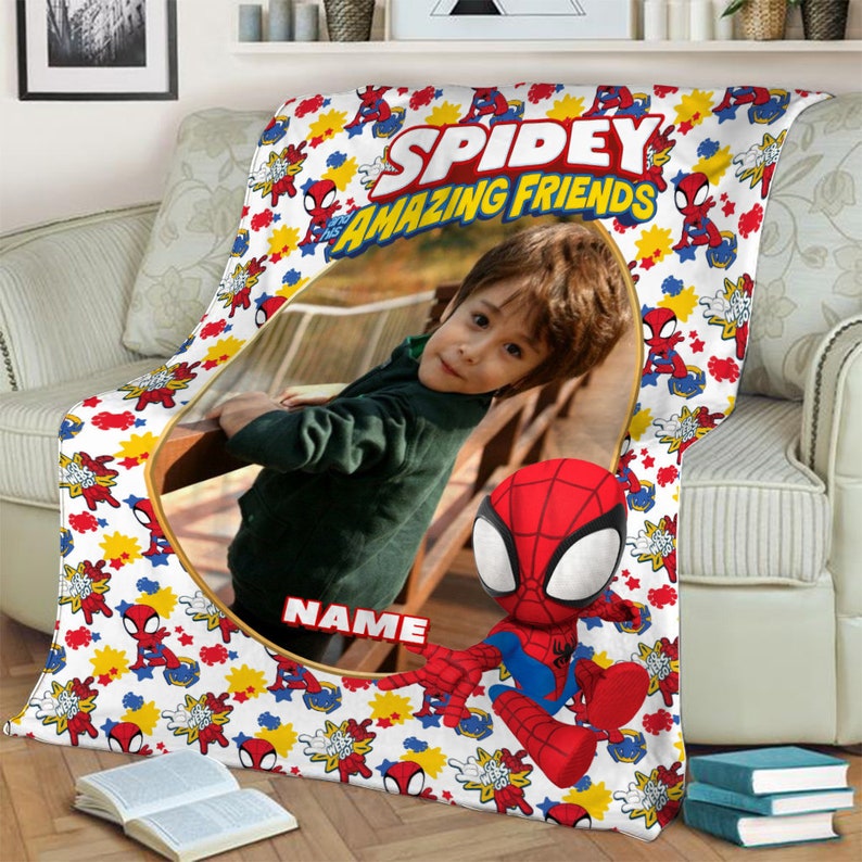 K&ouml;nnte beinhalten: Eine wei&szlig;e Fleecedecke mit einem wiederkehrenden Muster aus Spidey and His Amazing Friends-Figuren, gelben Sternen und roten Netzgrafiken. In der Mitte befindet sich ein gro&szlig;er ovaler Rahmen mit einem Kinderfoto und dem Wort NAME.
