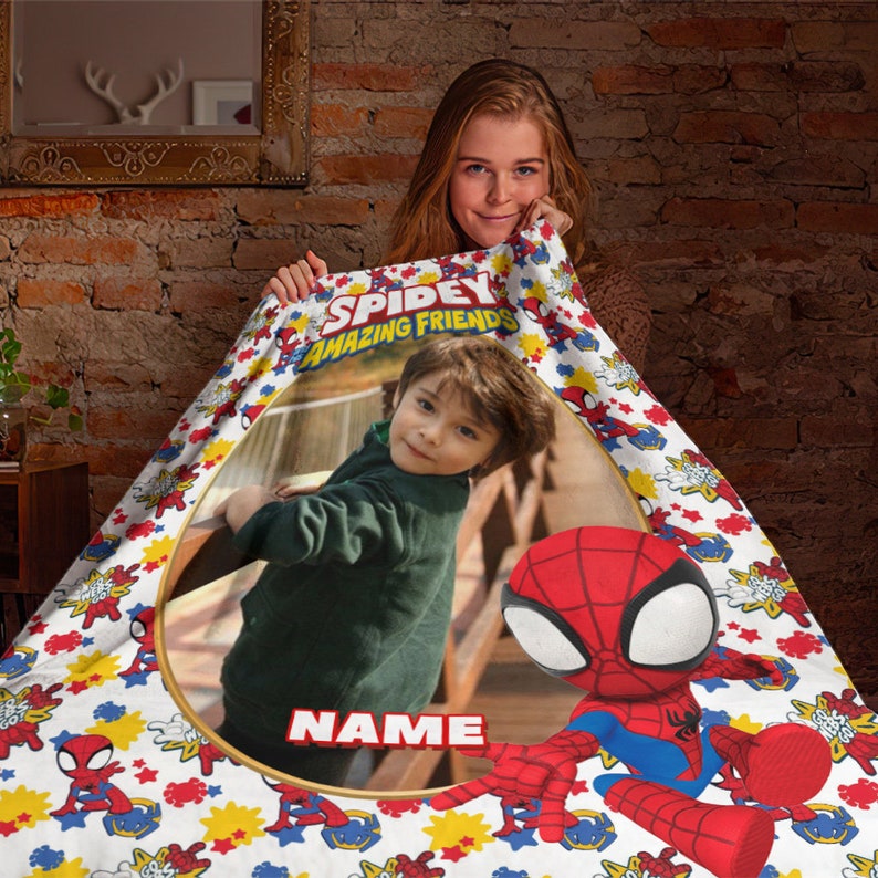 K&ouml;nnte beinhalten: Eine wei&szlig;e Fleecedecke mit einem wiederkehrenden Muster aus Spidey und seine Super-Freunde-Figuren, Sternen und Blitzen. Die Mitte zeigt ein kreisf&ouml;rmiges Foto eines Kindes in einer gr&uuml;nen Jacke mit dem Text Spidey Amazing Friends und Name.