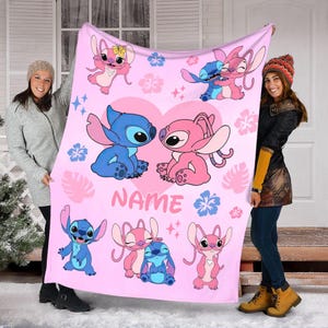 May include: A pink blanket featuring cartoon characters in blue and pink, with a heart design and floral accents. The blanket has the word "NAME" printed on it.