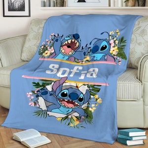 May include: A light blue fleece blanket featuring the character Stitch from Lilo and Stitch. The design includes tropical flowers, green leaves, and the name Sofia printed in white letters between two horizontal pink lines on the fabric.