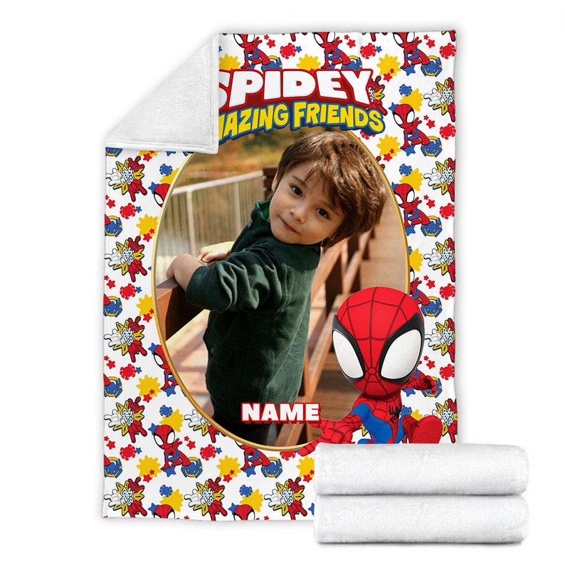 K&ouml;nnte beinhalten: Eine wei&szlig;e Fleecedecke mit einem sich wiederholenden Muster aus roten und blauen Spidey and His Amazing Friends-Figuren, gelben Sternen und roten Explosionen. In der Mitte befindet sich ein gro&szlig;er ovaler Fotorahmen mit dem Text Spidey and His Amazing Friends.