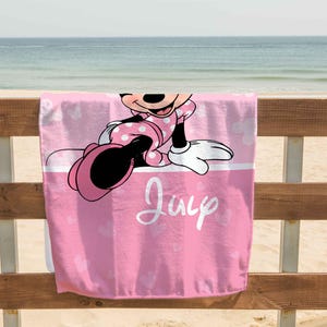 Puede incluir: Una toalla de playa rosa con un gr&aacute;fico de Minnie Mouse vistiendo un vestido de lunares. La toalla tiene una franja horizontal blanca en el medio con el nombre July escrito en cursiva blanca y un patr&oacute;n de corazones de color rosa claro.