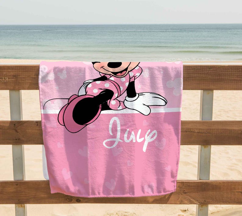 Puede incluir: Una toalla de playa rosa con un gr&aacute;fico de Minnie Mouse vistiendo un vestido de lunares. La toalla tiene una franja horizontal blanca en el medio con el nombre July escrito en cursiva blanca y un patr&oacute;n de corazones de color rosa claro.