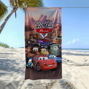 May include: A brown beach towel featuring a colorful illustration of characters from the movie "Cars." The design includes a red car with a smiling face, other animated cars, and the text "Name" on a red sign with checkered flags.