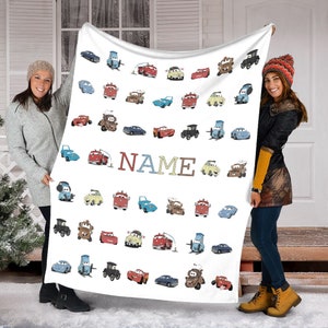 K&ouml;nnte beinhalten: Eine wei&szlig;e Fleecedecke mit einem sich wiederholenden Muster aus verschiedenen Zeichentrickautos aus dem Film Cars, darunter rote, blaue, gelbe und braune Fahrzeuge. Das Wort NAME ist mittig in gro&szlig;en, bunten Blockbuchstaben aufgedruckt.