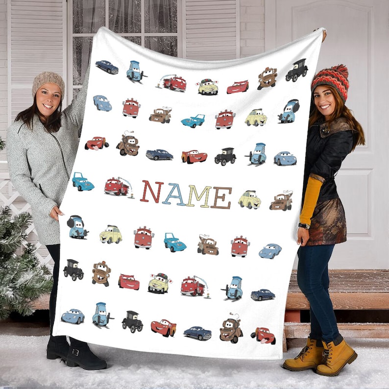 K&ouml;nnte beinhalten: Eine wei&szlig;e Fleecedecke mit einem sich wiederholenden Muster aus verschiedenen Zeichentrickautos aus dem Film Cars, darunter rote, blaue, gelbe und braune Fahrzeuge. Das Wort NAME ist mittig in gro&szlig;en, bunten Blockbuchstaben aufgedruckt.