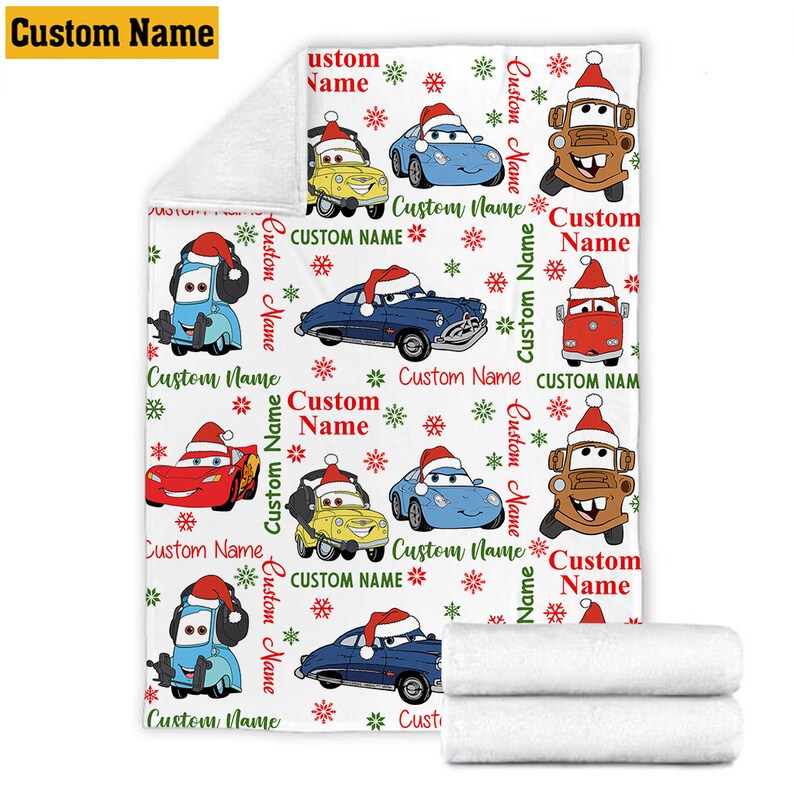 Puede incluir: Una manta de forro polar blanca con un patr&oacute;n repetitivo de coches de dibujos animados con gorros de Pap&aacute; Noel, intercalados con copos de nieve rojos y verdes y el texto Custom Name impreso en varias orientaciones. La manta tiene una esquina doblada.