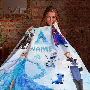 May include: A white fleece blanket with a watercolor design featuring characters from the movie Frozen. The blanket features Elsa, Anna, Olaf, Kristoff, and Sven. The blanket also features the text "NAME" in blue.