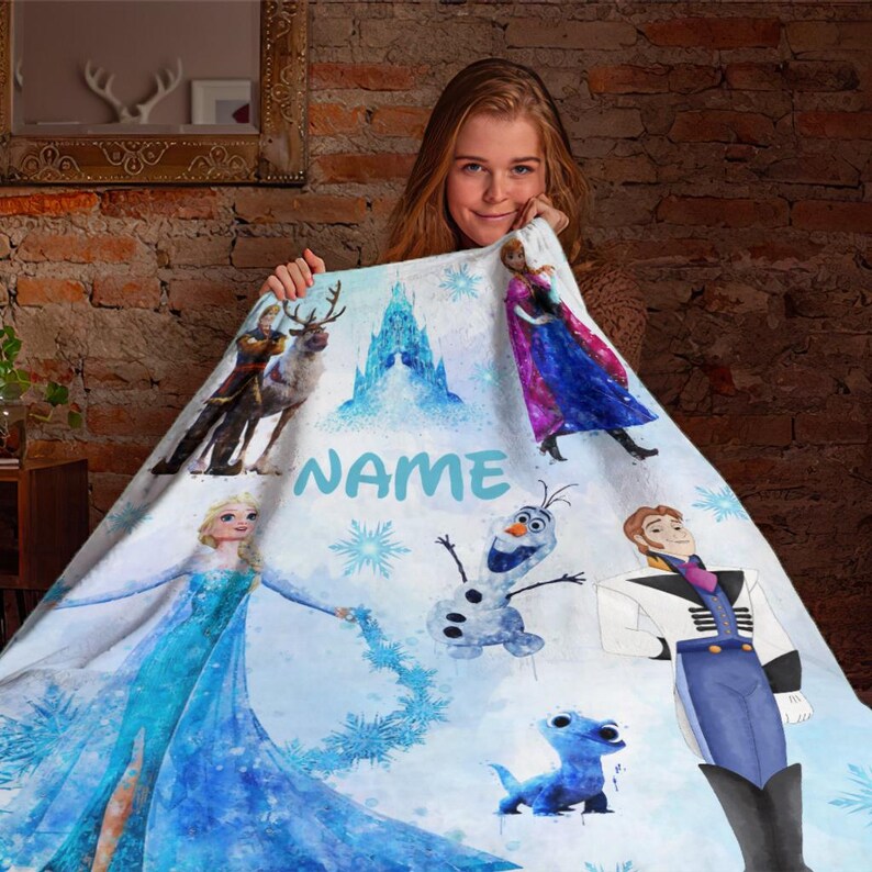 May include: A white fleece blanket with a watercolor design featuring characters from the movie Frozen. The blanket features Elsa, Anna, Olaf, Kristoff, and Sven. The blanket also features the text "NAME" in blue.