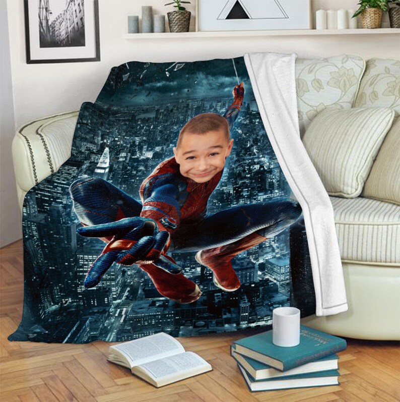 Puede incluir: Una manta de forro polar con una imagen impresa de un superh&eacute;roe balance&aacute;ndose por un horizonte urbano. El superh&eacute;roe lleva un traje rojo y azul con un logotipo de ara&ntilde;a en el pecho. La cara de un ni&ntilde;o se superpone a la cabeza del superh&eacute;roe.