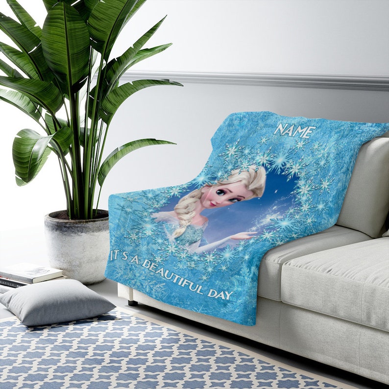 Pu&ograve; includere: Una coperta in pile azzurra con la grafica di un personaggio biondo con una treccia, circondato da un bordo di fiocchi di neve. Il testo "NAME" appare in alto e la frase "IT'S A BEAUTIFUL DAY" &egrave; stampata nella parte inferiore.