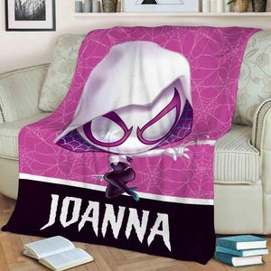 K&ouml;nnte beinhalten: Eine rosa und schwarze Fleece-Decke mit einem Cartoon-Bild einer wei&szlig;en Spinnenfrau mit einer lila Maske. Die Decke hat ein Spinnennetz-Design und der Name "JOANNA" ist unten aufgedruckt.