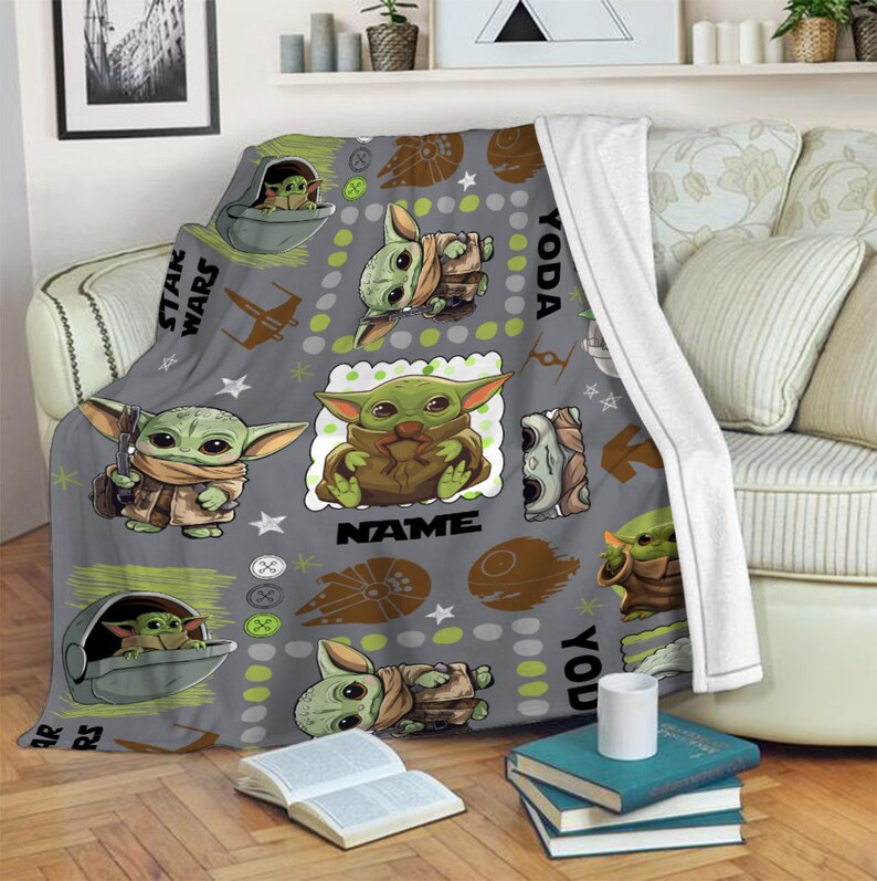 May include: A gray fleece throw blanket featuring a repeating pattern of the character Grogu from Star Wars, including illustrations of the character in a pod, the Millennium Falcon, and the Death Star, with the text Star Wars, Yoda, and NAME.