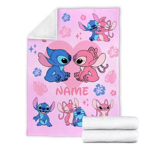 May include: Pink blanket featuring cartoon characters Stitch and Angel, with a heart and floral design. The blanket has a soft, plush texture and includes the word "NAME". Two white rolled towels are at the bottom.