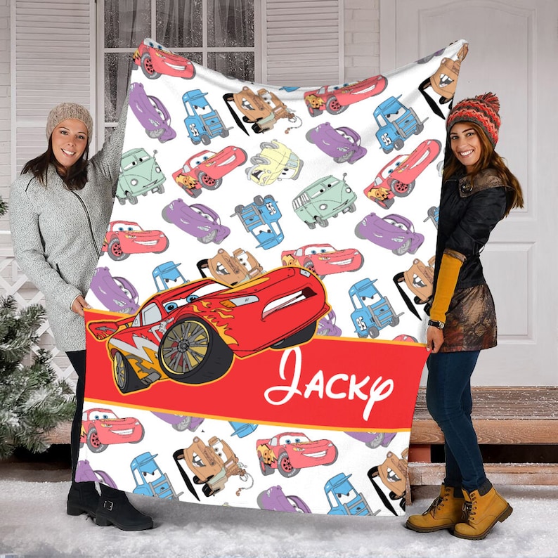 Pu&ograve; includere: Una coperta in pile bianca con un motivo ripetuto di varie auto da corsa a cartoni animati in rosso, blu, viola e marrone chiaro. Una grande auto da corsa rossa &egrave; mostrata sopra una fascia rossa con il testo corsivo bianco Jacky.