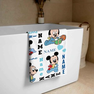May include: A white bath towel with a blue and white pattern featuring Mickey Mouse characters. The towel has the text "NAME" repeated throughout the design.