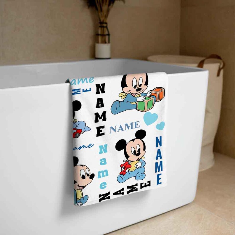 May include: A white bath towel with a blue and white pattern featuring Mickey Mouse characters. The towel has the text "NAME" repeated throughout the design.