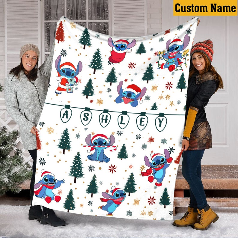 May include: A white fleece blanket featuring a repeating pattern of the character Stitch wearing Santa hats, interspersed with green pine trees, gold snowflakes, and a string of lights spelling the name ASHLEY in dark green capital letters.