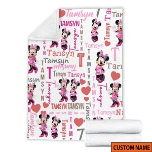 May include: A white fleece blanket featuring a repeating pattern of Minnie Mouse in a pink polka dot dress, red hearts, and the name Tamsyn printed in various pink, red, and grey fonts. The blanket has a folded corner and a rolled section at the bottom.