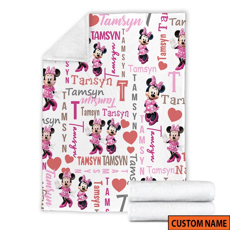 May include: A white fleece blanket featuring a repeating pattern of Minnie Mouse in a pink polka dot dress, red hearts, and the name Tamsyn printed in various pink, red, and grey fonts. The blanket has a folded corner and a rolled section at the bottom.