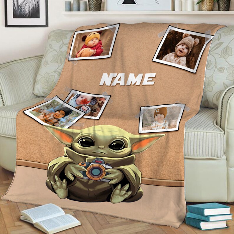 May include: A tan fleece blanket with a brown corkboard background. The blanket features a cartoon image of a green baby Yoda holding a camera. There are four photo frames with pictures of babies on the corkboard. The text "NAME" is printed on the corkboard.