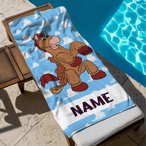 May include: A blue beach towel with a cartoon horse character in brown and red, set against a sky-blue backdrop with white clouds. The towel has a white stripe with the word "NAME" in dark purple.