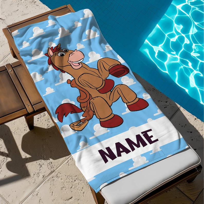 May include: A blue beach towel with a cartoon horse character in brown and red, set against a sky-blue backdrop with white clouds. The towel has a white stripe with the word "NAME" in dark purple.