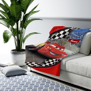 May include: A soft throw blanket featuring a red race car character with blue eyes and a smiling mouth. The design includes a black and white checkered border, a stadium crowd background, and a red banner with the word Name printed in white.