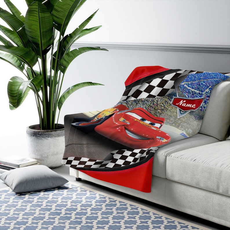 May include: A soft throw blanket featuring a red race car character with blue eyes and a smiling mouth. The design includes a black and white checkered border, a stadium crowd background, and a red banner with the word Name printed in white.