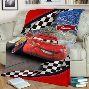 May include: A soft throw blanket featuring a red race car with blue eyes and a smiling mouth, set against a background of a racetrack and a crowd. The design includes black and white checkered borders and a red emblem with the text Name.