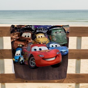 May include: A beach towel featuring a colorful graphic of animated cars. The design includes a red race car, a blue car, a yellow car, a green van, and other vehicles. The towel is displayed on a wooden fence with a beach and ocean in the background.