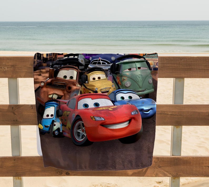 May include: A beach towel featuring a colorful graphic of animated cars. The design includes a red race car, a blue car, a yellow car, a green van, and other vehicles. The towel is displayed on a wooden fence with a beach and ocean in the background.