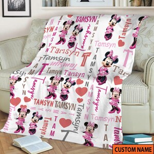 May include: A white fleece blanket featuring a repeating pattern of Minnie Mouse in a pink polka dot dress, pink hearts, and the name Tamsyn printed in various pink, red, and grey fonts. The design includes the letter T in a large block style.