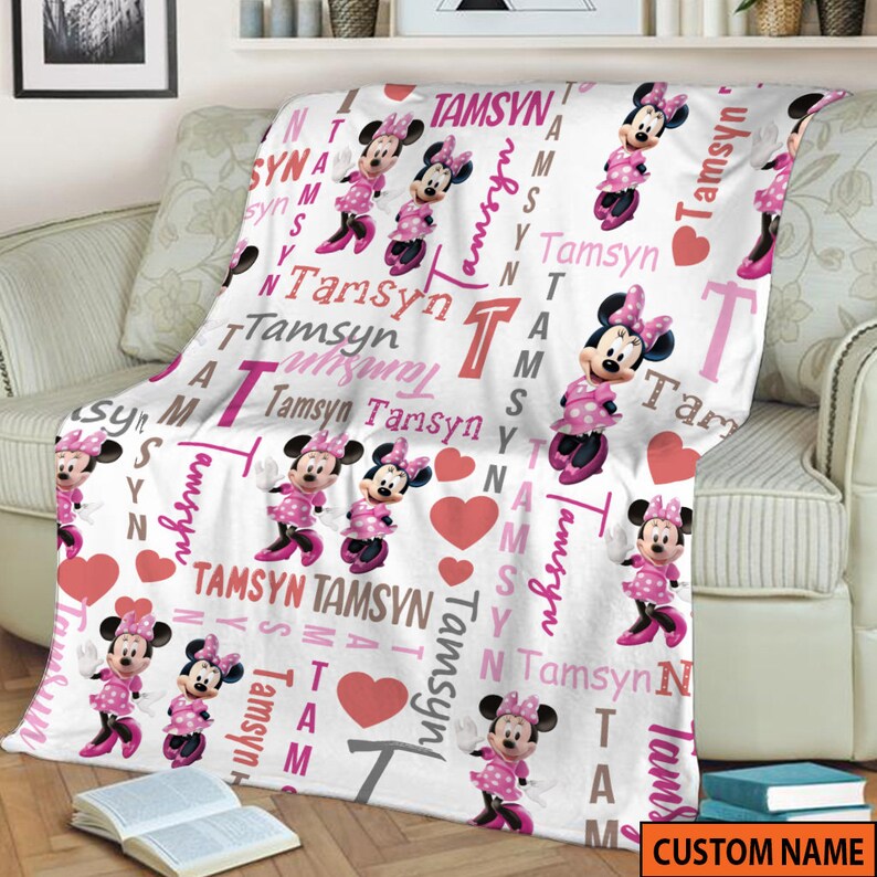 May include: A white fleece blanket featuring a repeating pattern of Minnie Mouse in a pink polka dot dress, pink hearts, and the name Tamsyn printed in various pink, red, and grey fonts. The design includes the letter T in a large block style.