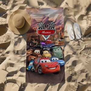 May include: A rectangular beach towel featuring characters from the animated movie "Cars." The towel has a brown background with images of race cars, the text "Name," and checkered flags. A straw hat and white flip-flops are on the sand.